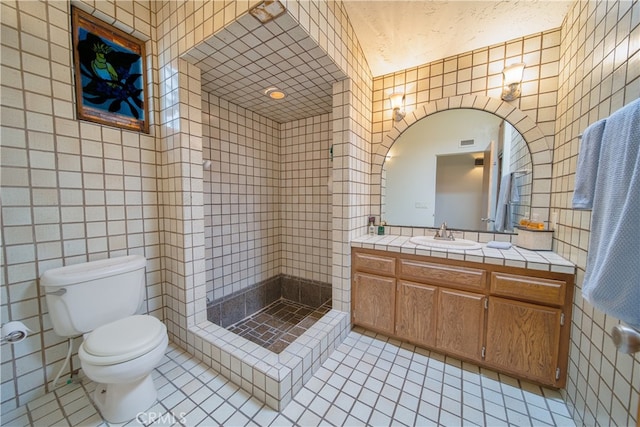 bathroom with a tile shower, tile walls, tile patterned flooring, vanity, and toilet