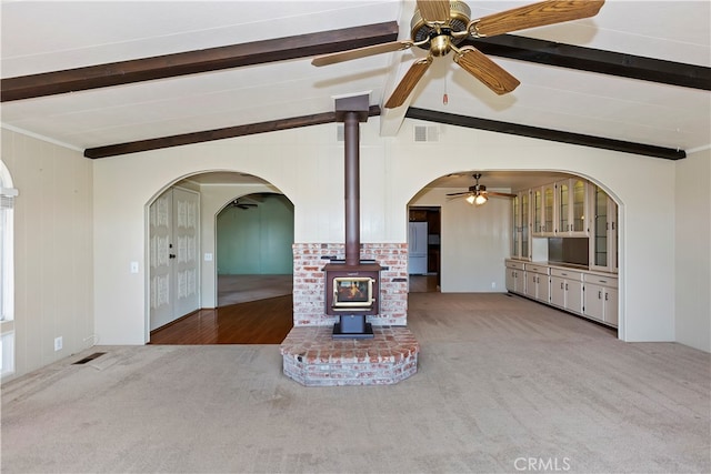 unfurnished living room with ceiling fan, vaulted ceiling with beams, light colored carpet, and a wood stove