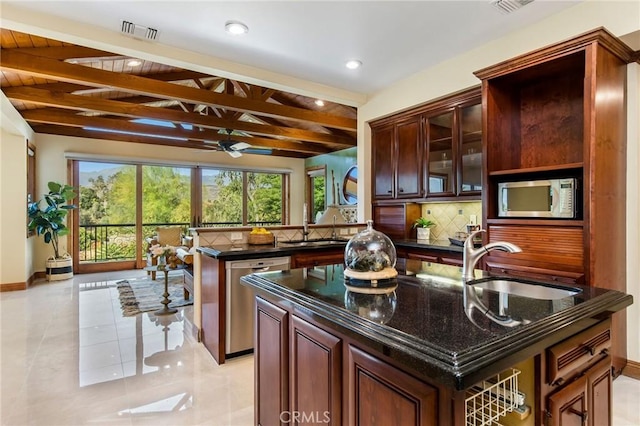 kitchen featuring stainless steel appliances, beamed ceiling, sink, backsplash, and a center island with sink