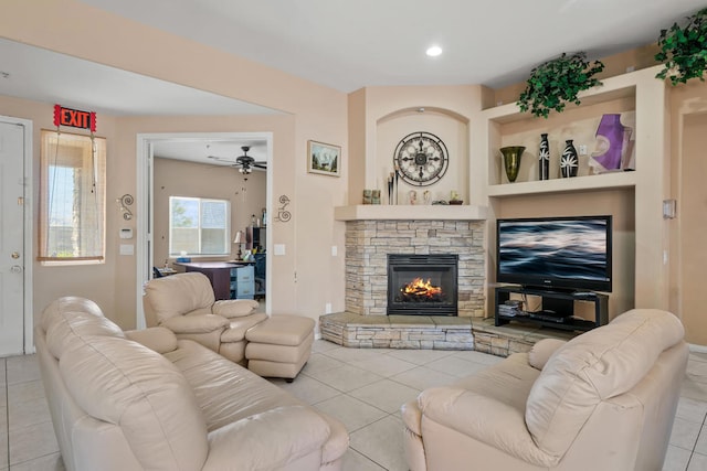 tiled living room with ceiling fan, built in features, and a stone fireplace