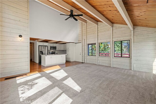 unfurnished living room with high vaulted ceiling, wood walls, light wood-type flooring, beam ceiling, and ceiling fan