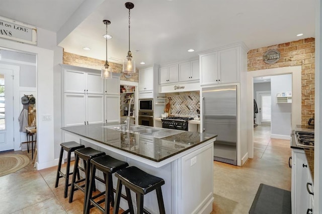 kitchen featuring a breakfast bar area, built in appliances, a center island with sink, pendant lighting, and white cabinetry