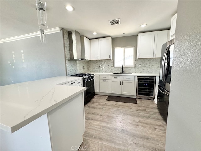 kitchen featuring white cabinetry, kitchen peninsula, beverage cooler, black gas stove, and wall chimney range hood