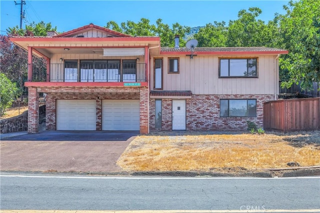 view of front facade featuring a balcony, aphalt driveway, an attached garage, fence, and brick siding