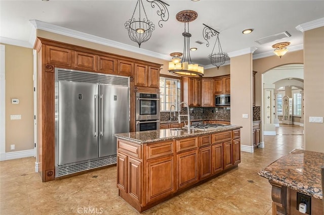 kitchen featuring dark stone counters, stainless steel appliances, crown molding, decorative light fixtures, and an island with sink