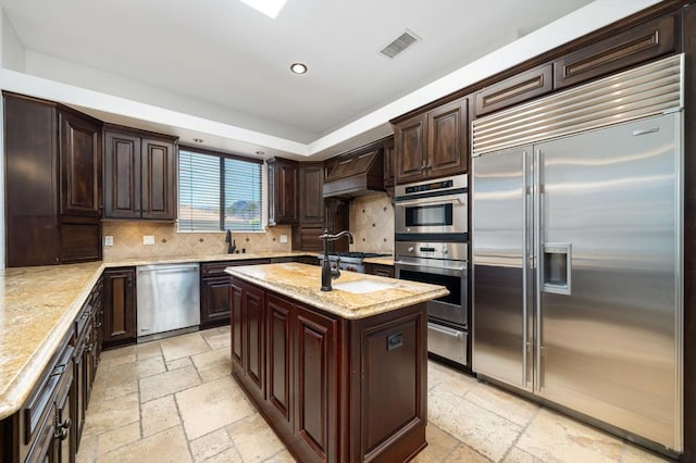 kitchen featuring a center island with sink, backsplash, premium range hood, and stainless steel appliances