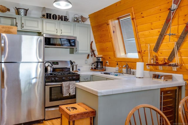 kitchen with tile counters, sink, kitchen peninsula, wooden walls, and appliances with stainless steel finishes