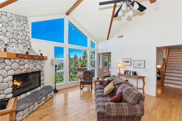 living room with light hardwood / wood-style floors, beam ceiling, high vaulted ceiling, a stone fireplace, and ceiling fan