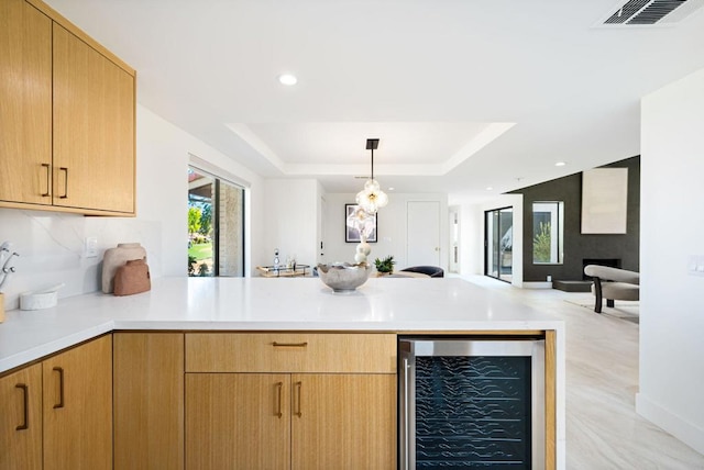 kitchen with a raised ceiling, kitchen peninsula, decorative light fixtures, wine cooler, and light brown cabinets