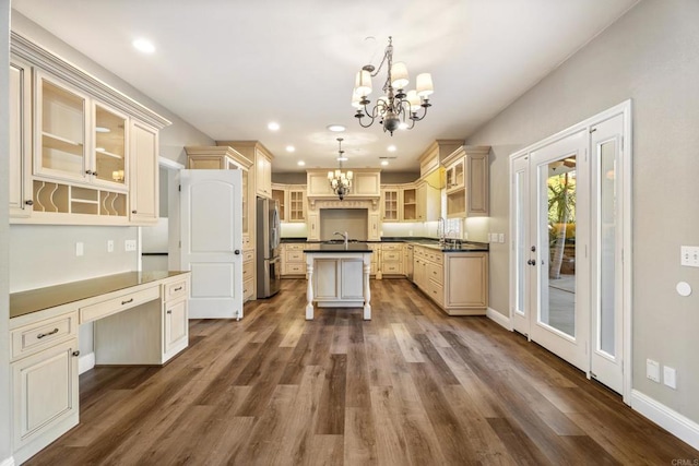 kitchen with dark wood-type flooring, cream cabinets, a center island with sink, hanging light fixtures, and stainless steel fridge