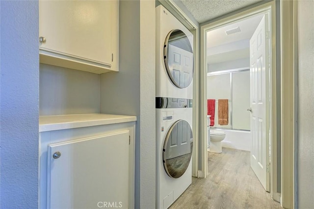 clothes washing area featuring light wood-type flooring, visible vents, a textured ceiling, stacked washing maching and dryer, and cabinet space