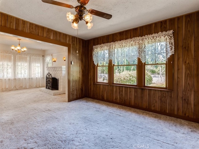 unfurnished living room with wood walls, a brick fireplace, a textured ceiling, carpet, and ceiling fan with notable chandelier