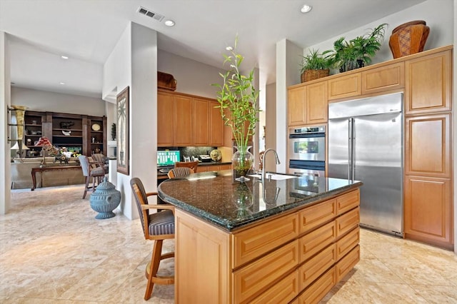 kitchen with appliances with stainless steel finishes, dark stone counters, a kitchen island with sink, sink, and a breakfast bar area