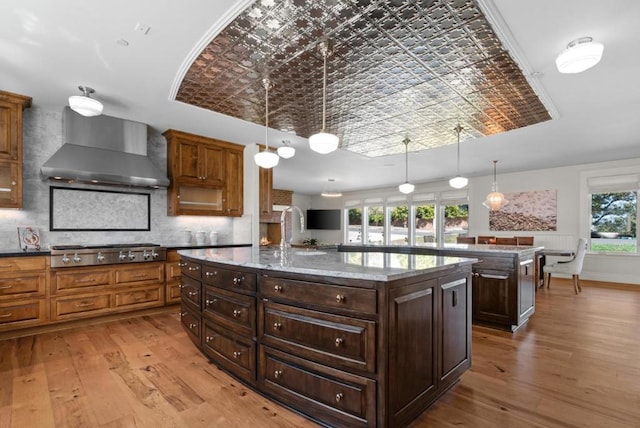 kitchen with wall chimney exhaust hood, hanging light fixtures, stainless steel gas stovetop, a kitchen island with sink, and light wood-type flooring