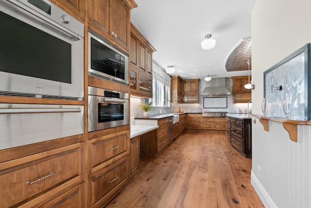 kitchen featuring backsplash, wall chimney range hood, hanging light fixtures, appliances with stainless steel finishes, and light hardwood / wood-style floors