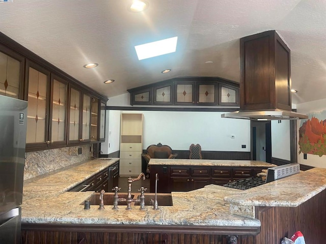 kitchen with stainless steel refrigerator, island range hood, kitchen peninsula, vaulted ceiling with skylight, and dark brown cabinets