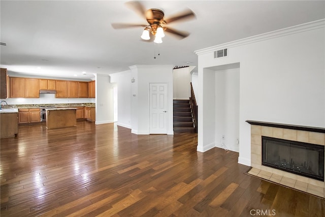 unfurnished living room featuring ceiling fan, ornamental molding, sink, a tile fireplace, and dark hardwood / wood-style flooring