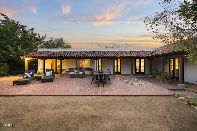 back of house at dusk with stucco siding, a tile roof, an outdoor living space, a patio, and french doors