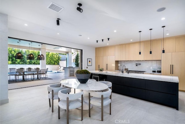 kitchen with a kitchen island with sink, hanging light fixtures, light tile patterned floors, light brown cabinetry, and tasteful backsplash