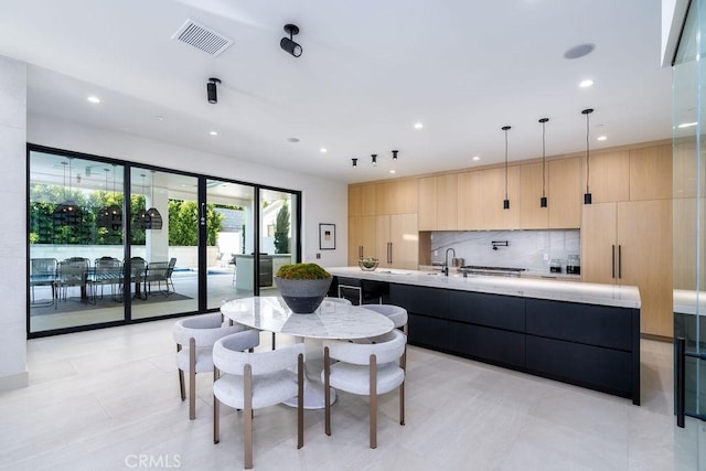 kitchen with light brown cabinets, backsplash, decorative light fixtures, a center island with sink, and light tile patterned flooring