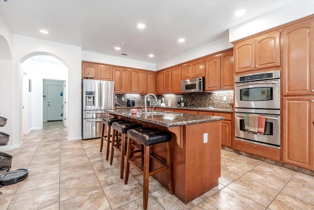 kitchen with sink, appliances with stainless steel finishes, dark stone countertops, an island with sink, and a kitchen bar