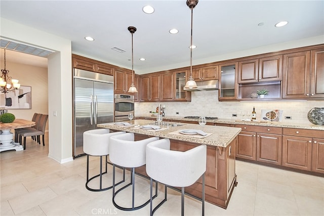 kitchen with light stone counters, hanging light fixtures, a kitchen island with sink, appliances with stainless steel finishes, and decorative backsplash