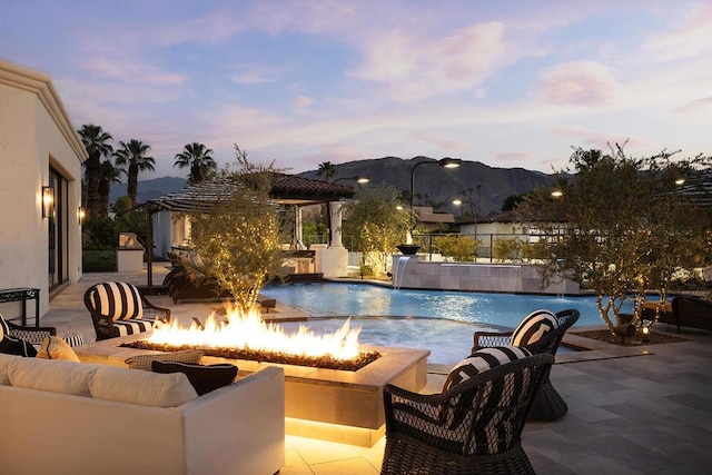 pool at dusk featuring a mountain view, pool water feature, a fire pit, and a patio area