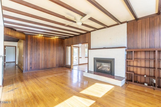 unfurnished living room featuring wood walls, a brick fireplace, ceiling fan, beamed ceiling, and wood-type flooring