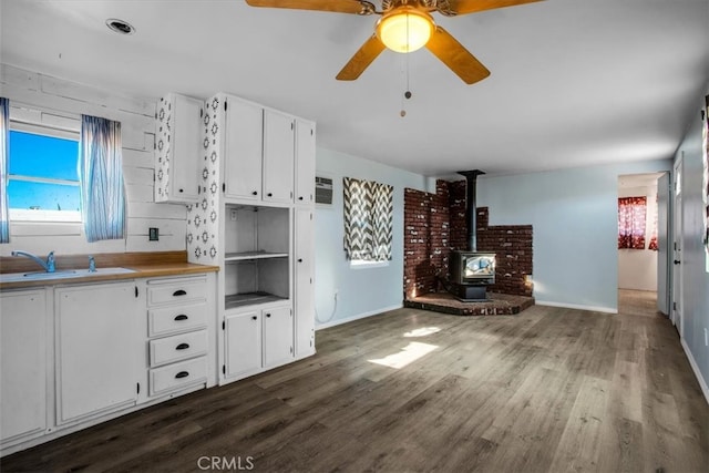 kitchen with white cabinetry, dark wood-type flooring, a wood stove, ceiling fan, and sink