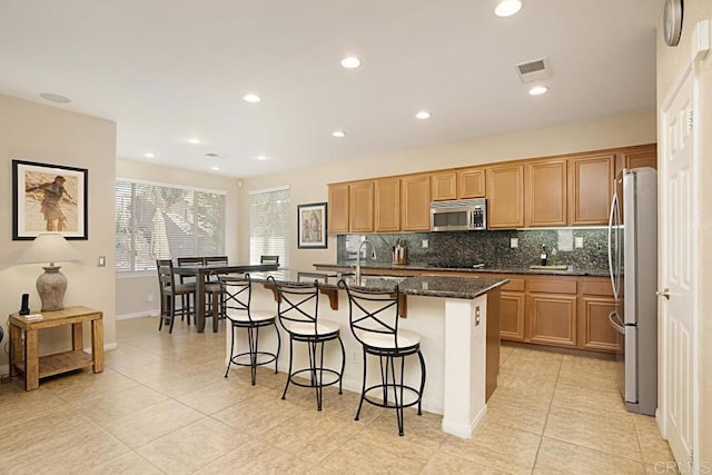 kitchen with backsplash, an island with sink, dark stone counters, light tile patterned floors, and appliances with stainless steel finishes