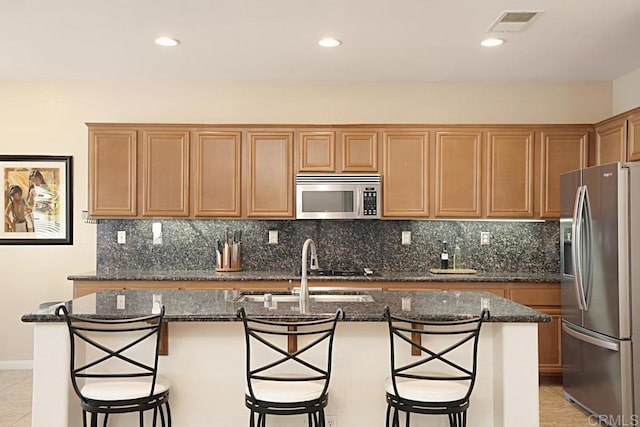 kitchen with appliances with stainless steel finishes, backsplash, a kitchen breakfast bar, dark stone counters, and an island with sink