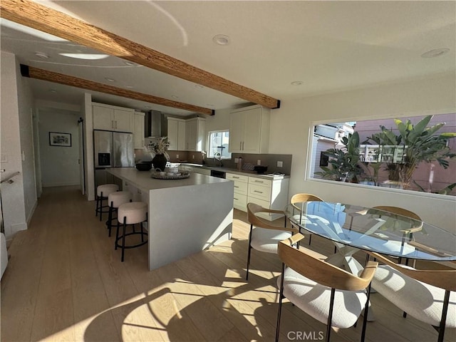 kitchen with light hardwood / wood-style flooring, wall chimney exhaust hood, stainless steel refrigerator with ice dispenser, white cabinets, and beamed ceiling