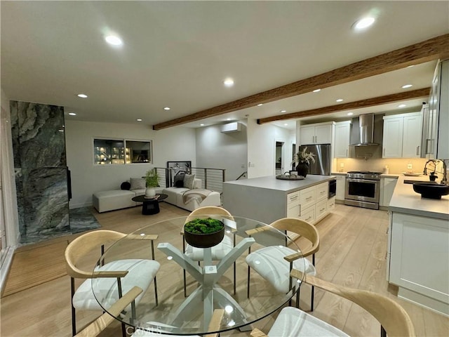 kitchen with wall chimney range hood, beam ceiling, stainless steel appliances, light hardwood / wood-style floors, and white cabinets