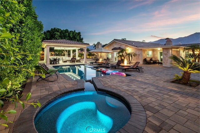 pool at dusk with a gazebo, an in ground hot tub, a mountain view, and a patio area