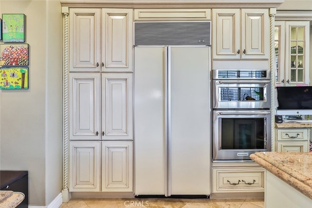 kitchen with light tile patterned flooring, light stone countertops, stainless steel double oven, built in fridge, and cream cabinets