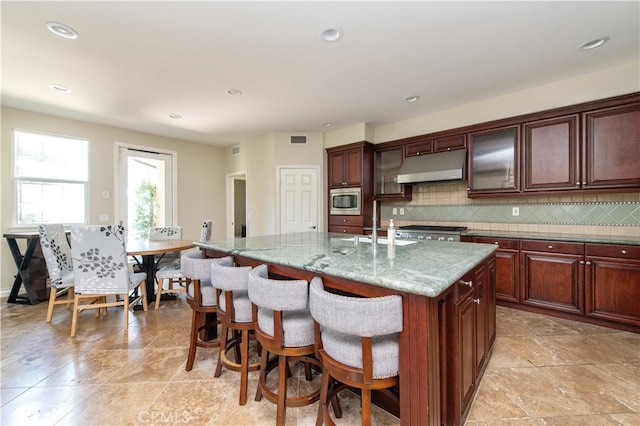 kitchen with a kitchen breakfast bar, decorative backsplash, an island with sink, light stone counters, and stainless steel microwave