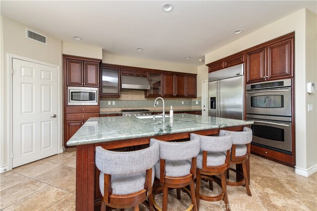 kitchen featuring light stone countertops, a center island with sink, built in appliances, and tasteful backsplash