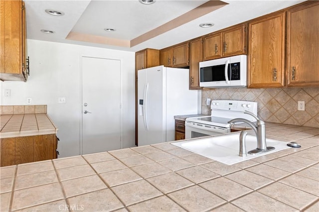 kitchen featuring tile counters, sink, backsplash, white appliances, and a tray ceiling