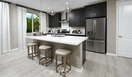 kitchen featuring a breakfast bar, stainless steel appliances, a kitchen island with sink, wall chimney range hood, and hanging light fixtures