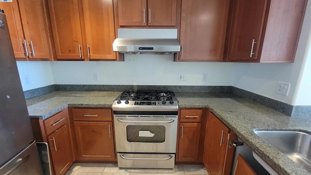 kitchen with range hood, stainless steel gas range, light tile patterned floors, dark stone countertops, and refrigerator