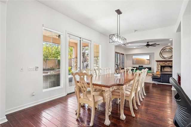 dining room with ceiling fan, a stone fireplace, and dark wood-type flooring