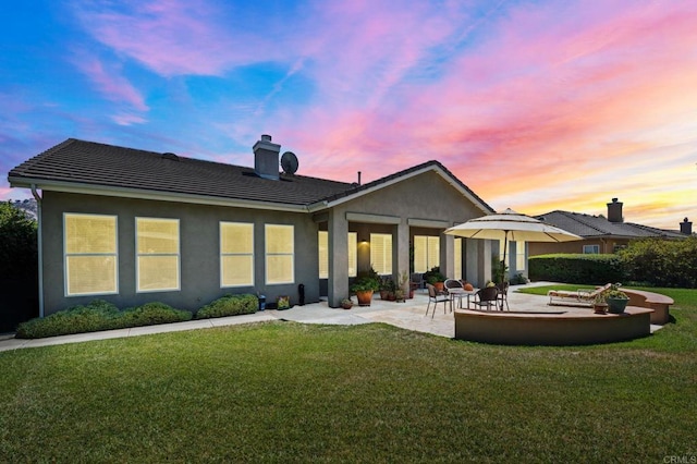 back of house at dusk featuring a patio, a chimney, a tiled roof, a yard, and stucco siding