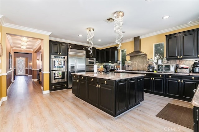 kitchen featuring appliances with stainless steel finishes, light wood-type flooring, ornamental molding, wall chimney exhaust hood, and a kitchen island