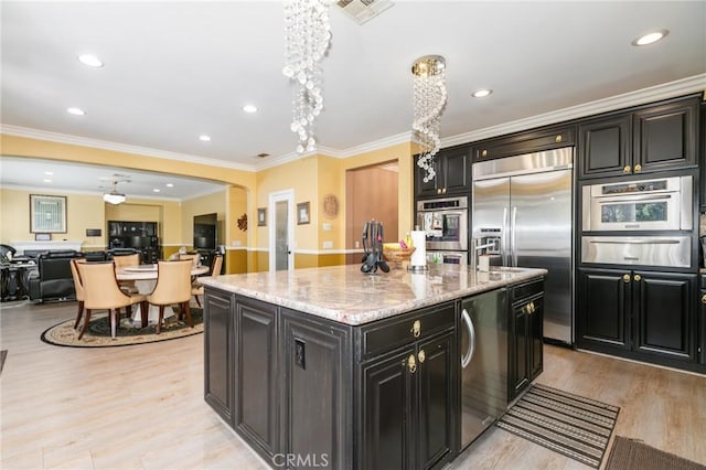 kitchen with light stone countertops, crown molding, an island with sink, appliances with stainless steel finishes, and light wood-type flooring