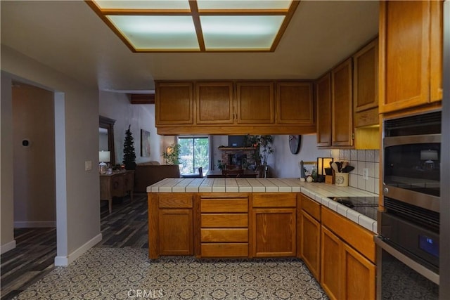 kitchen with stainless steel microwave, oven, decorative backsplash, tile counters, and kitchen peninsula