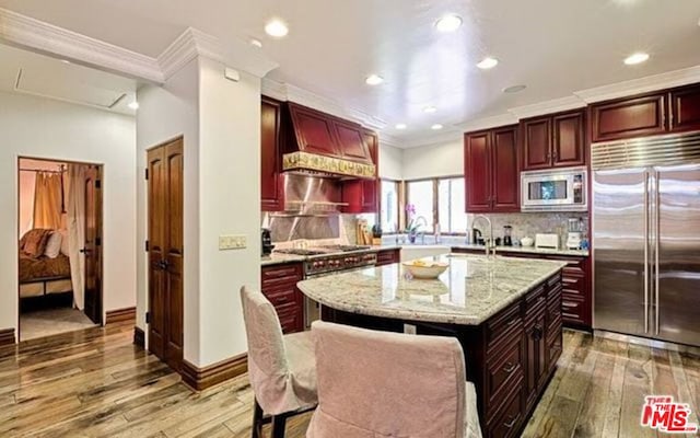 kitchen with custom exhaust hood, an island with sink, backsplash, built in appliances, and hardwood / wood-style flooring