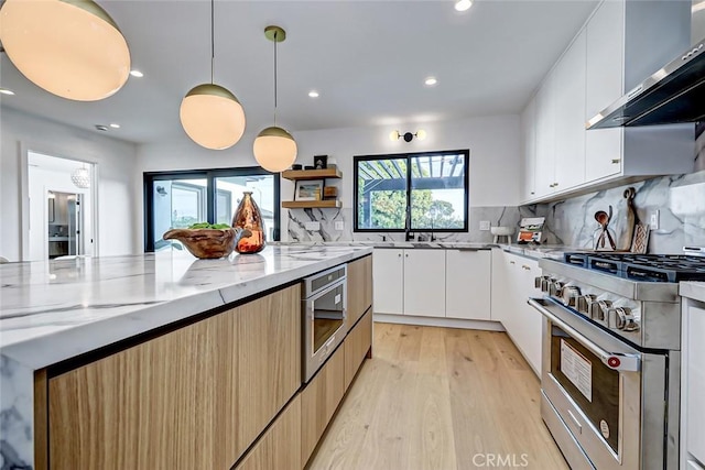 kitchen with decorative light fixtures, white cabinetry, light stone countertops, stainless steel appliances, and wall chimney exhaust hood