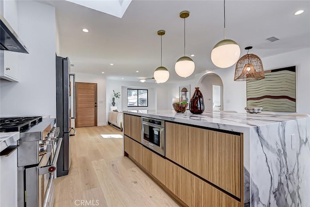 kitchen with wall chimney range hood, high end range, pendant lighting, light wood-type flooring, and light stone counters