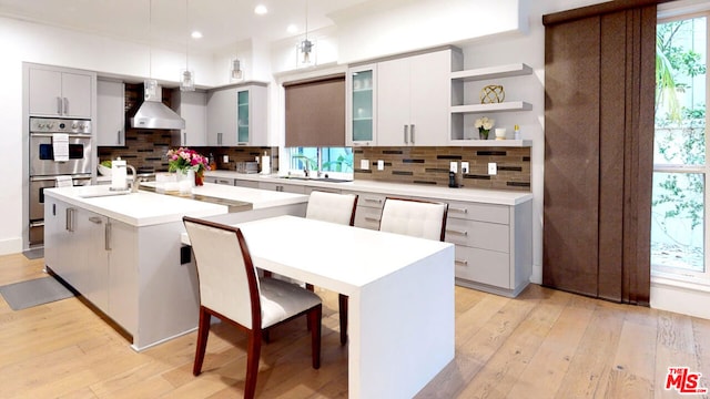 kitchen with an island with sink, pendant lighting, light hardwood / wood-style flooring, and wall chimney exhaust hood