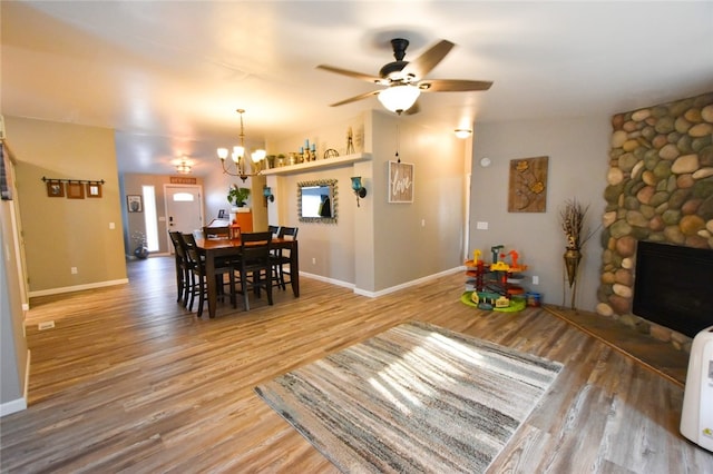 dining room featuring wood-type flooring, a stone fireplace, and ceiling fan with notable chandelier